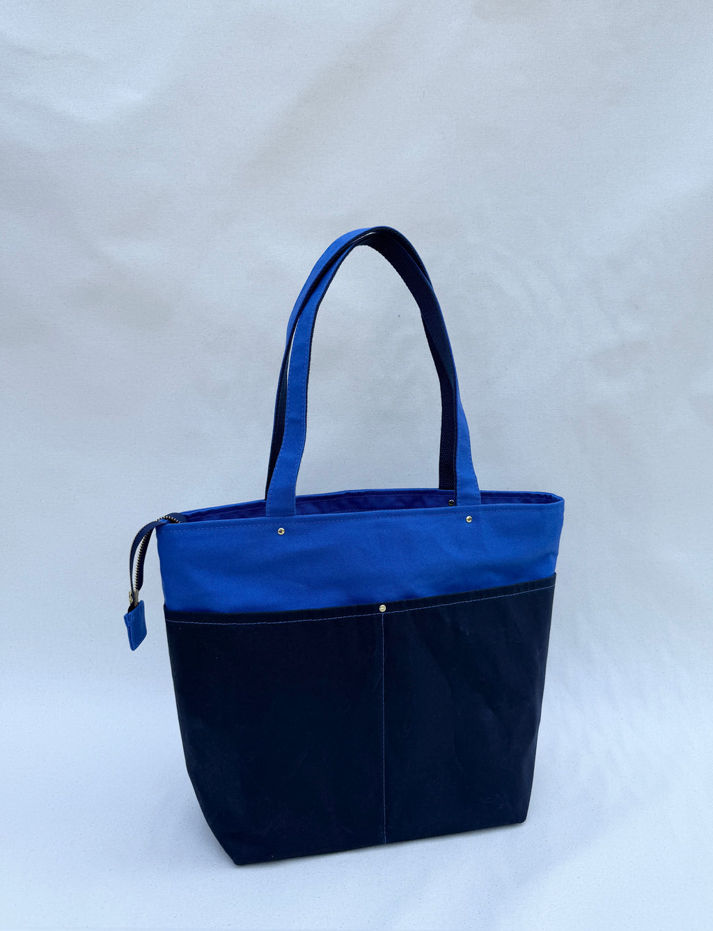 A royal blue canvas tote bag with dual shoulder straps, two front pockets in navy waxed canvas, and a top zipper, shown on a natural canvas backdrop