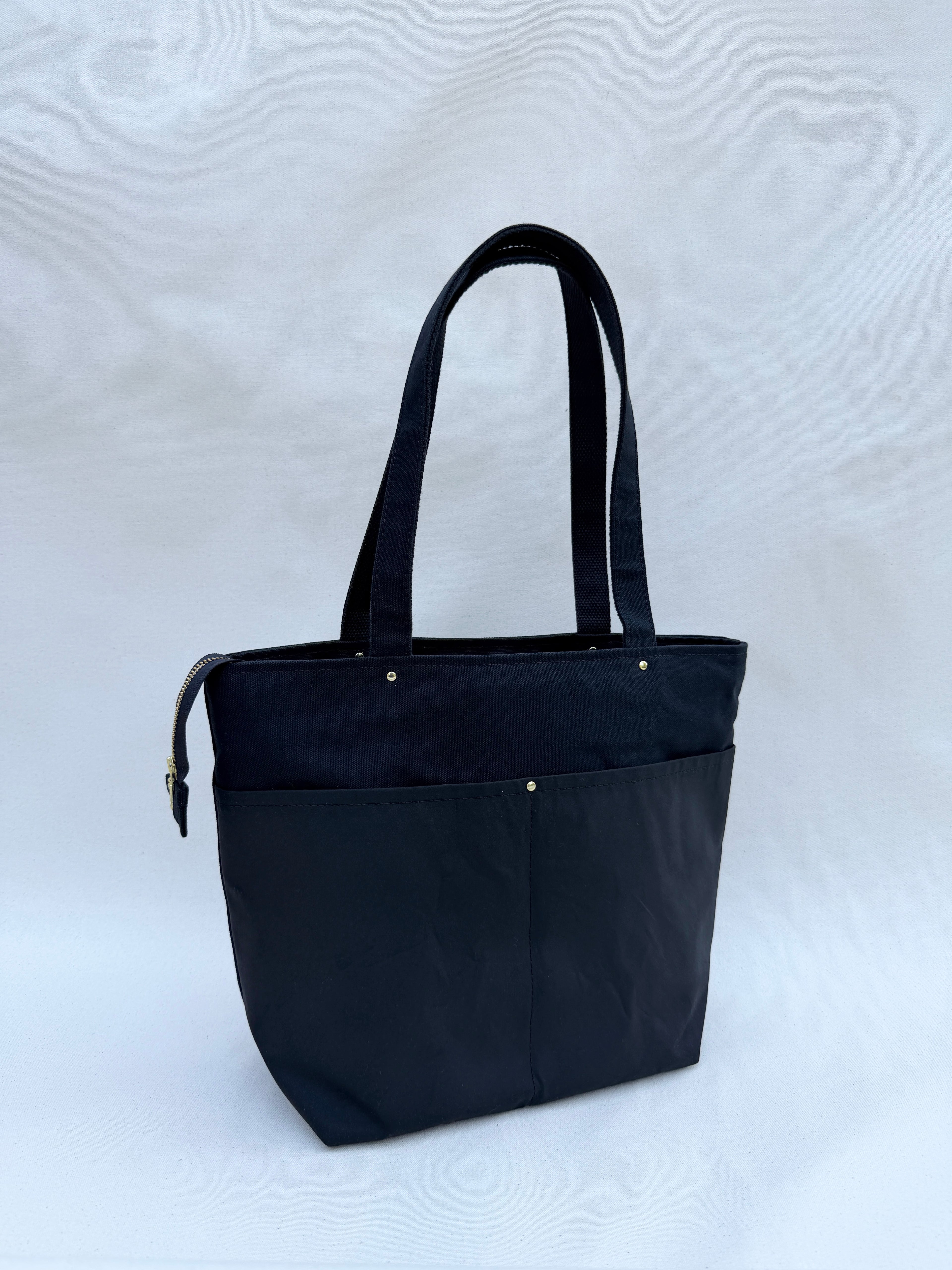 A black canvas bag with dual shoulder straps, two front pockets, and a top zipper, shown on a natural canvas backdrop
