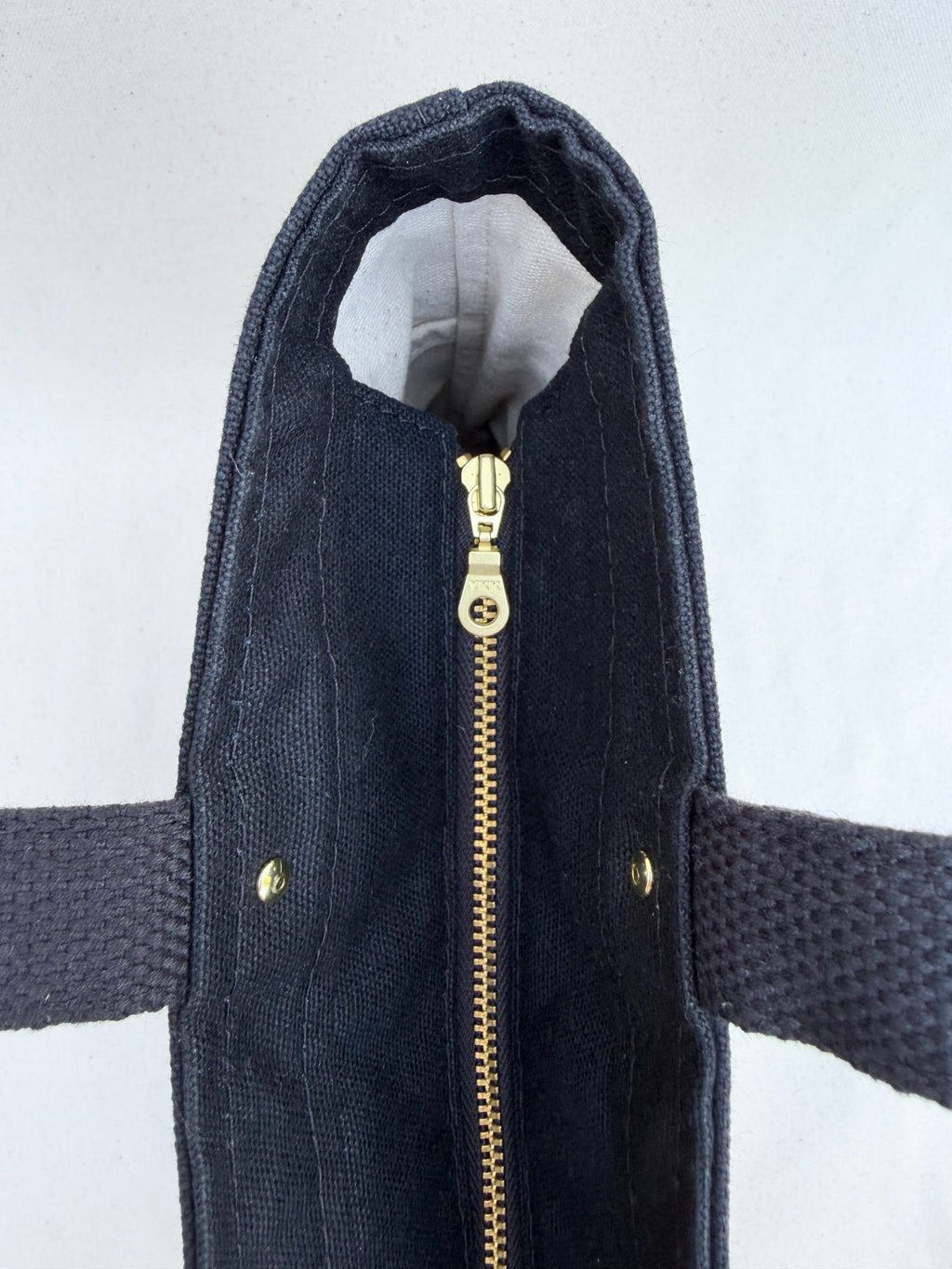 Close-up of a black canvas tote bag with a brass metal zipper and matching brass rivets, in front of a natural canvas backdrop