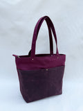 A deep purple-red tote bag with dual shoulder straps, two front pockets, and a top zipper, shown on a natural canvas backdrop