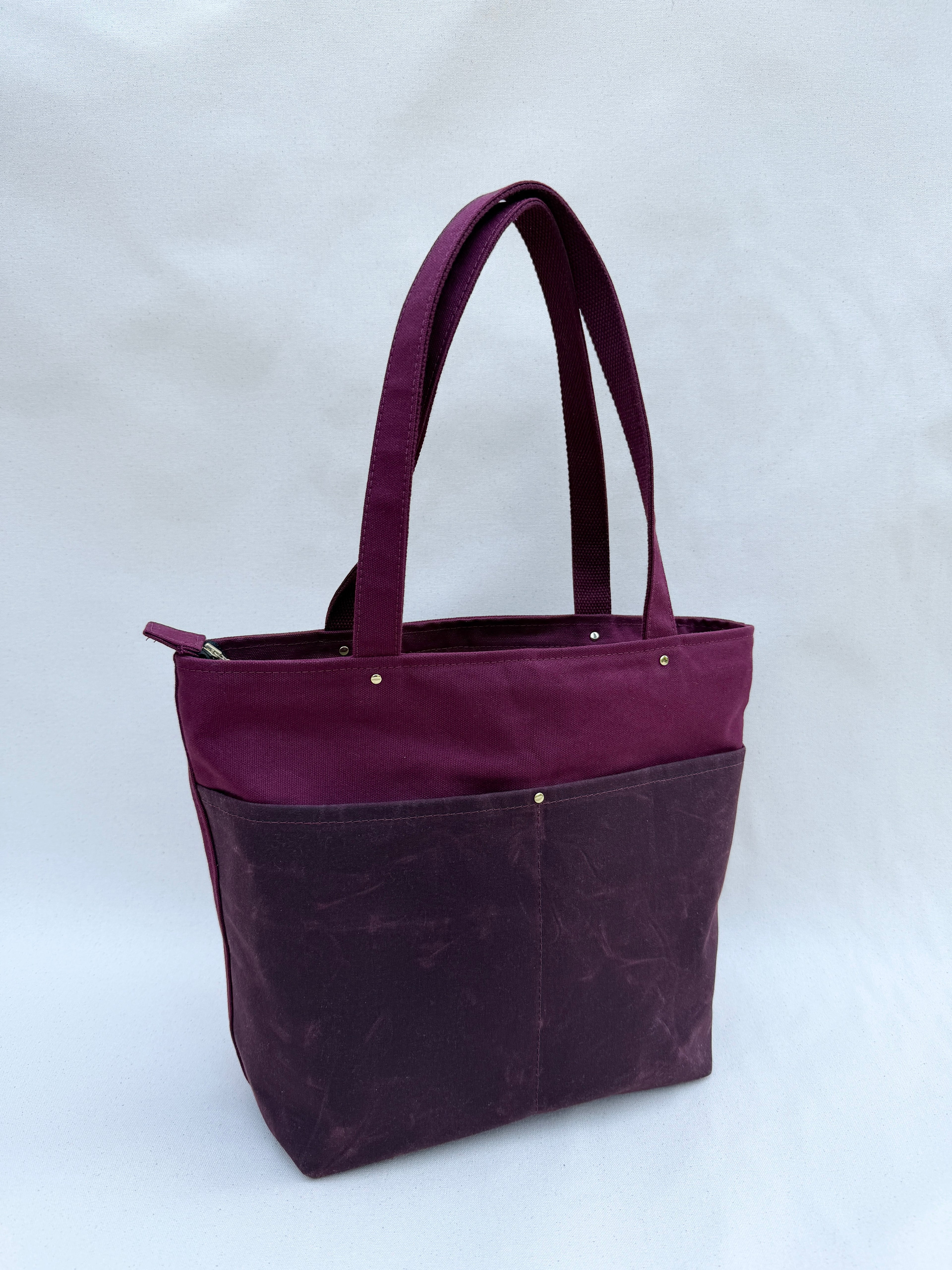 A deep purple-red tote bag with dual shoulder straps, two front pockets, and a top zipper, shown on a natural canvas backdrop
