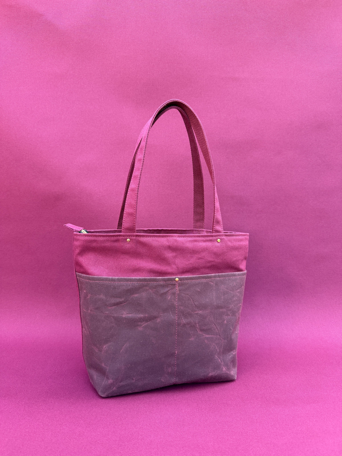 A maroon canvas bag with dual shoulder straps, two front pockets, and a top zipper, shown on a maroon canvas backdrop
