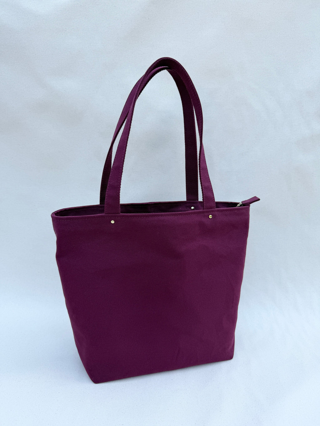 A deep reddish-purple tote with brass rivets in front of a natural canvas backdrop