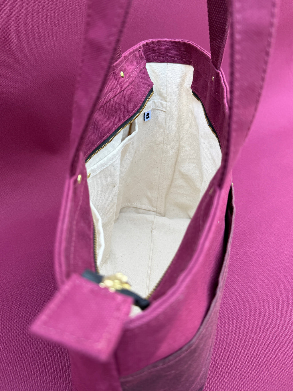 A close-up of a maroon tote bag with a natural canvas interior, showing the zipper opening and inner lining against a matching maroon background.