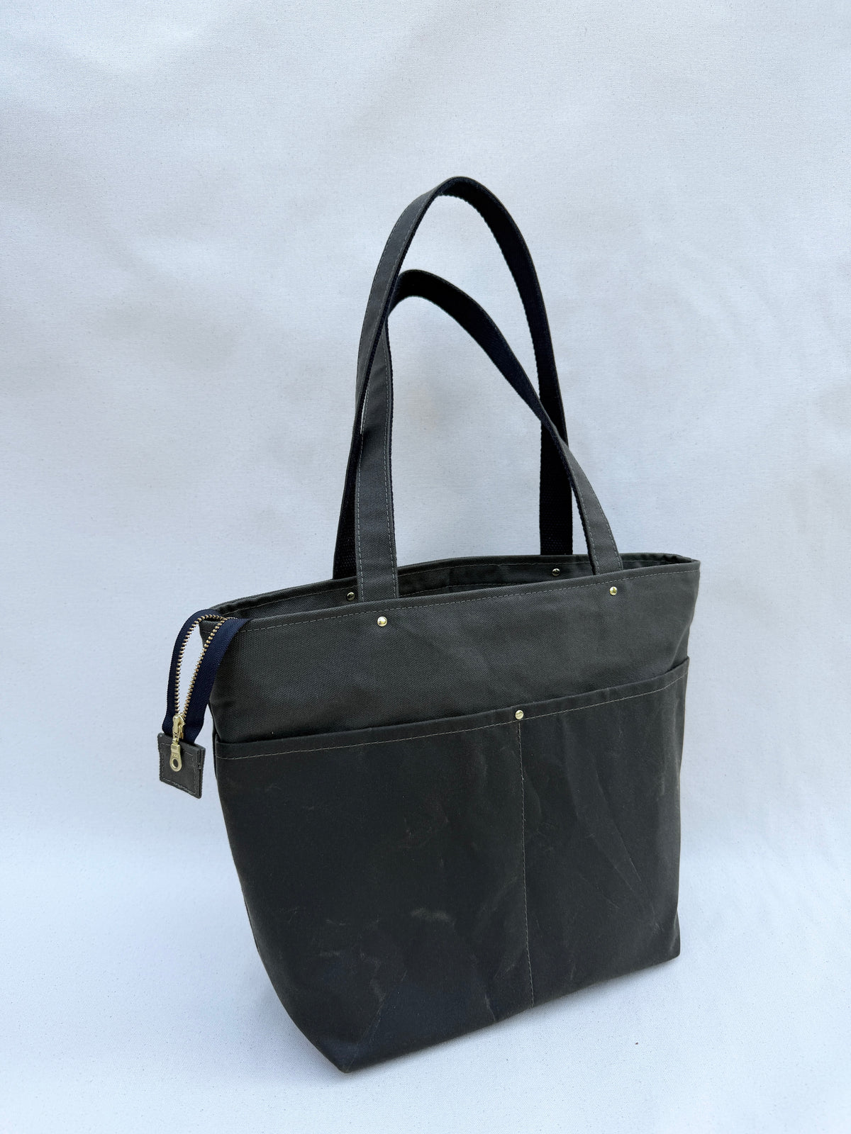 An olive drab green tote bag with dual shoulder straps, two front pocket, and a top zipper, shown on a natural canvas backdrop