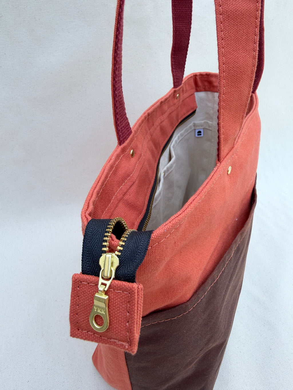A close-up of a canvas tote bag with a terracotta pink upper section, a dark brown bottom pocket, and a black zipper with a brass pull tab.
