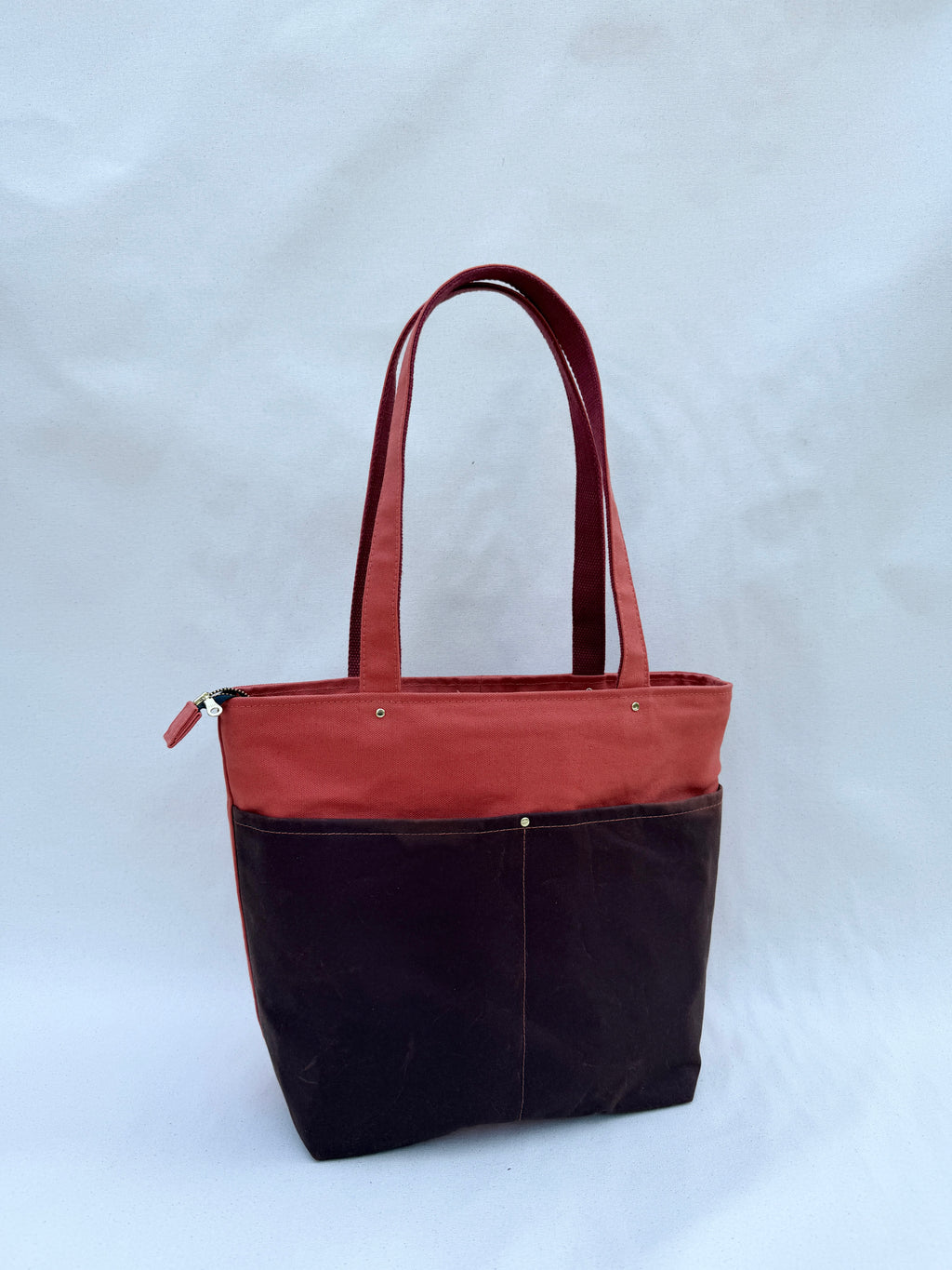 A tote bag with a terra cotta canvas top and handles, and a dark brown waxed canvas lower body, shown against a natural canvas backdrop.