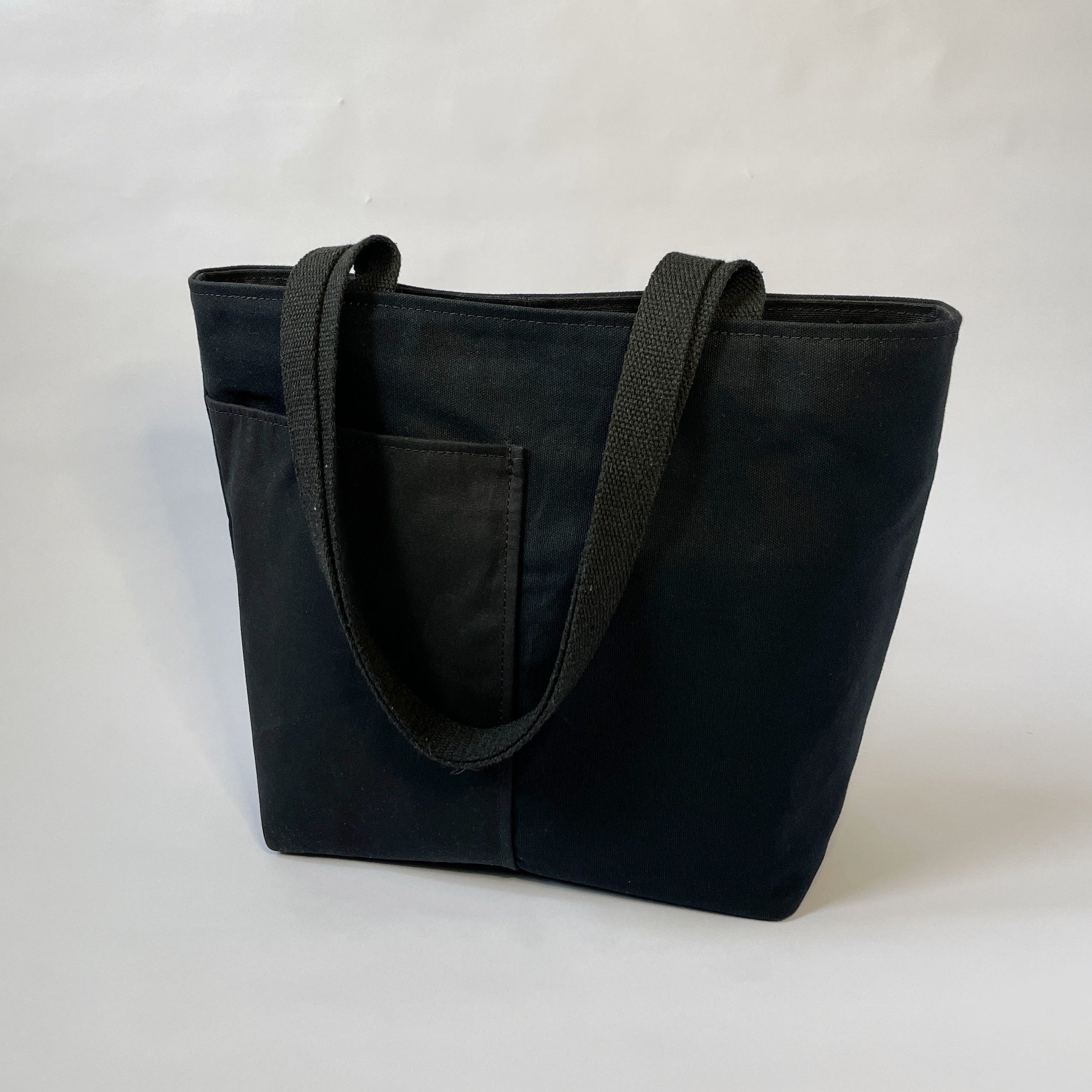 A black canvas tote bag with two handles, two lined exterior pockets, and two interior pockets, displayed against a neutral background.
