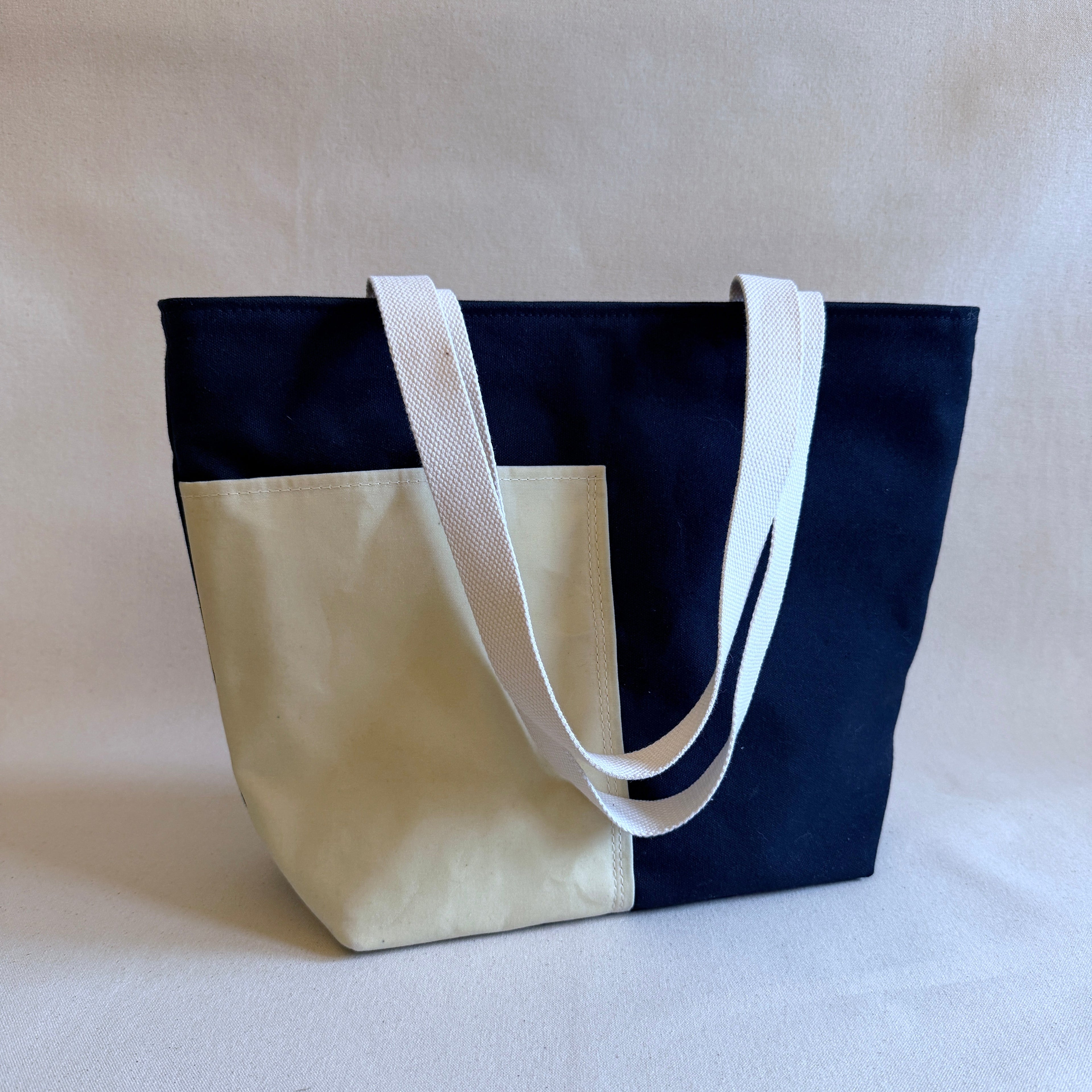 A black canvas tote bag with natural canvas exterior pockets and two natural canvas handles on a neutral backdrop.