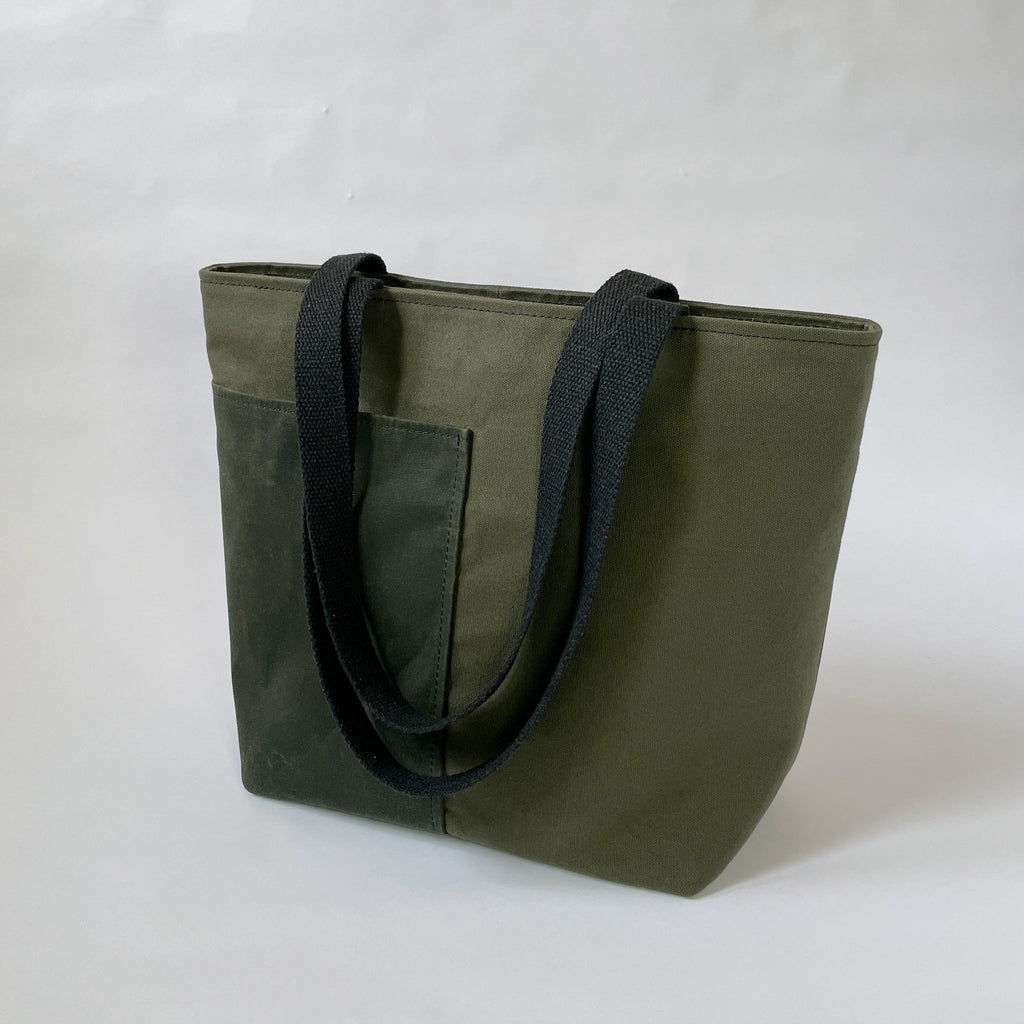 A dark olive canvas tote bag with two black handles, two lined exterior pockets, and two interior pockets, displayed against a neutral background.