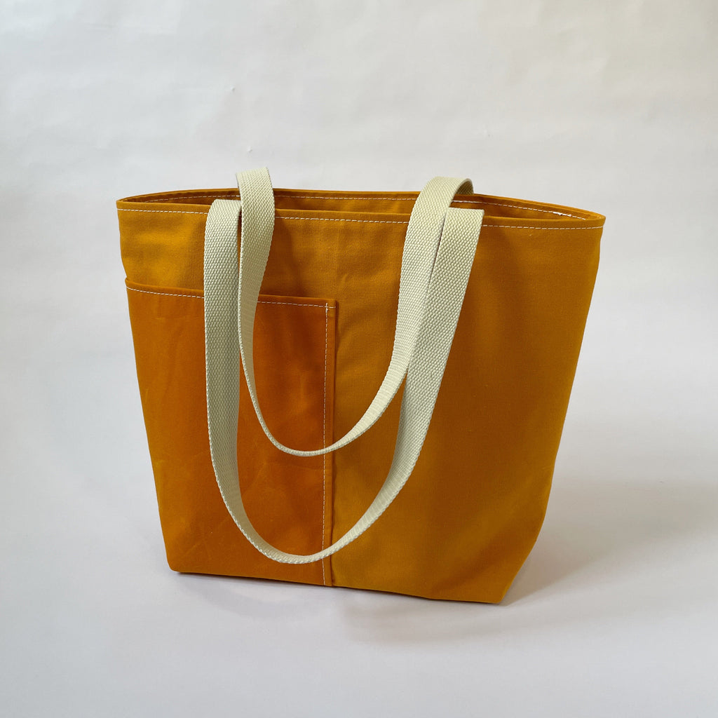 A yellow-orange canvas tote bag with two ivory handles, two lined exterior pockets, and two interior pockets, displayed against a neutral background.